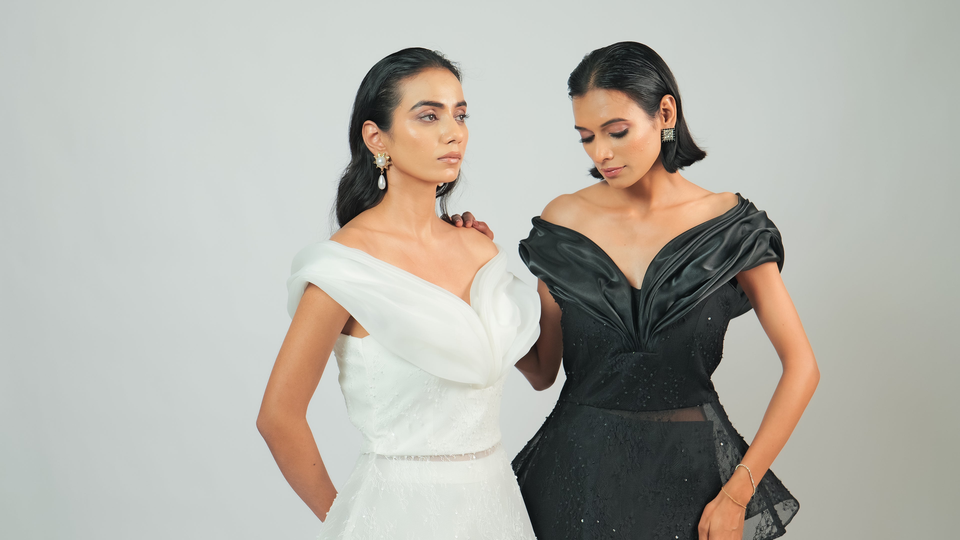 Two women wearing off-shoulder dresses, one white and one black, on a plain background.